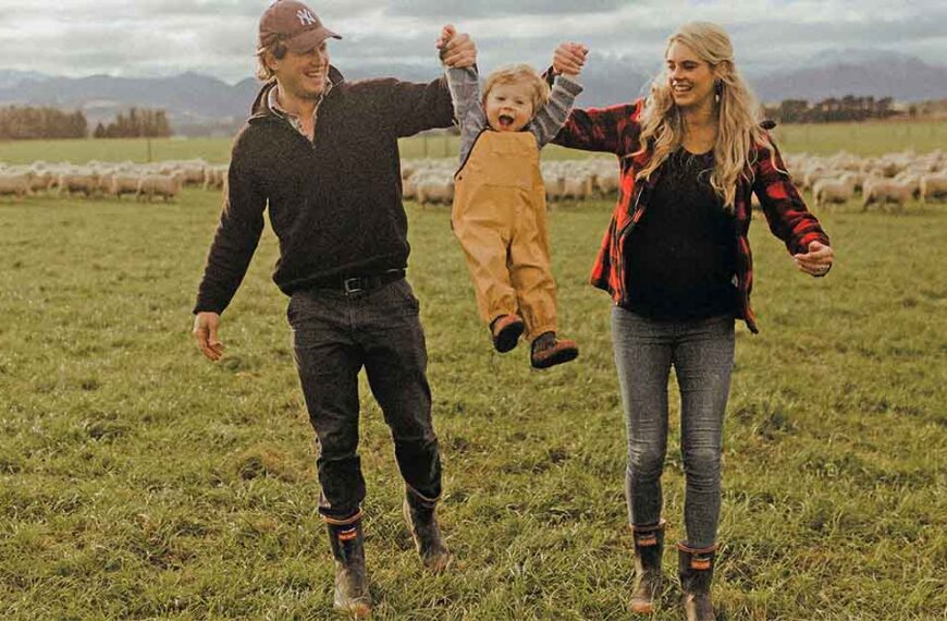 Angus and Elise Aitken lift up their young son George. They are standing in a paddock with sheep behind them.