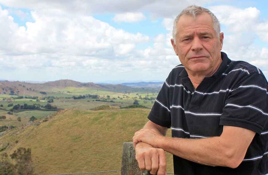 Farmer Doug Dobbs in front of the Waikato valley where he farms. It is greenish hill country with a few trees dotted about.
