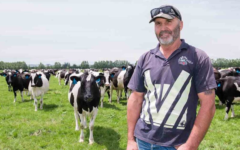 John Van Hout stands in a paddock on his dairy farm with a herd of black and white cows behind him.