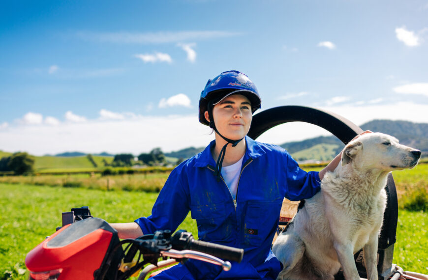 farm worker wearing a blue overall sits in a two-wheel-drive with a working farm dog at their side.