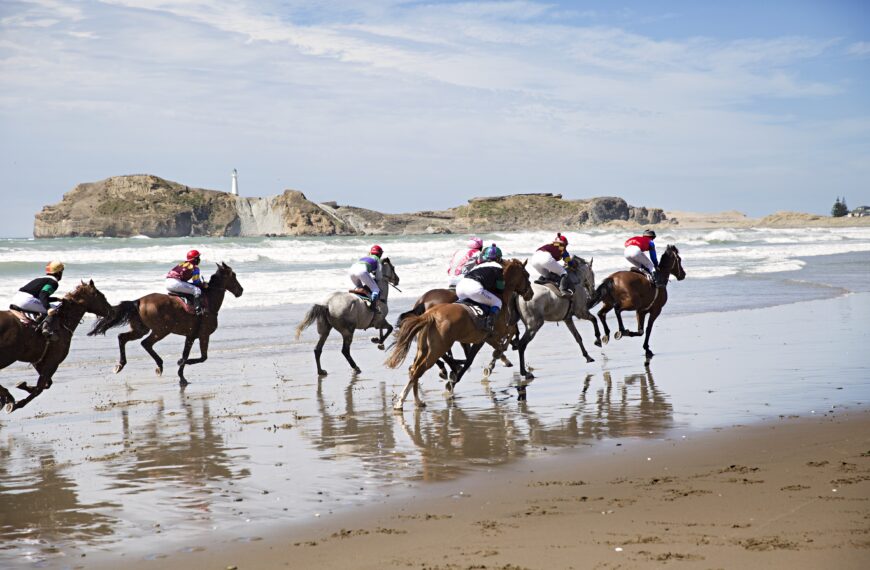A group of riders on horseback gallop along the beach shore.