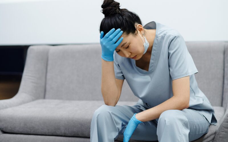 A tired nurse rests on a couch with her head in her hands.