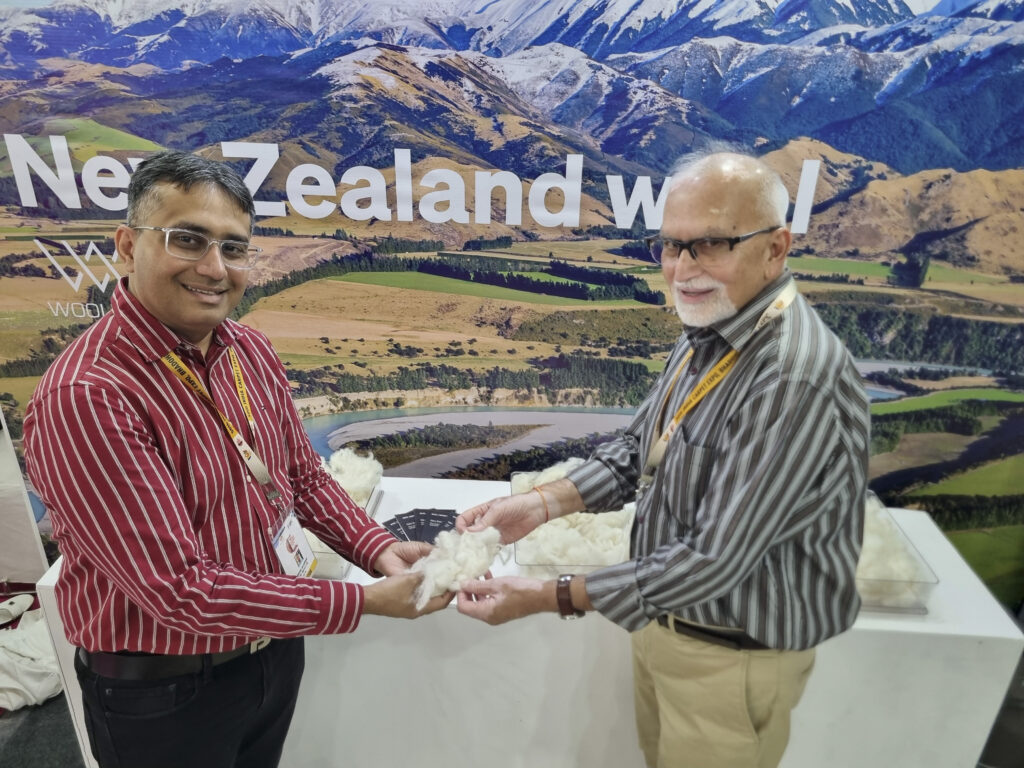 | Farmers Weekly Two men in striped shirts exchange wool at a booth with a New Zealand wool backdrop, Haas on lanyards and fluffy white wool on the table behind them.